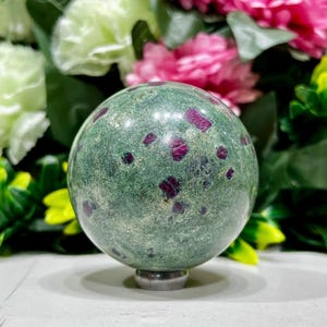 May include: A green sphere with red flecks, possibly a gemstone or decorative object. The sphere is resting on a silver base, with a blurred floral background of pink and green. The sphere's surface has a mottled appearance.