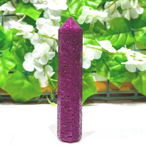 Può includere: Un alto obelisco di cristallo magenta scuro, sfaccettato, è centrato su uno sfondo di foglie verdi e fiori bianchi. Il cristallo ha una superficie strutturata e una parte superiore appuntita. L'obelisco è un oggetto decorativo.