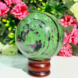 May include: A green sphere with dark green and purple inclusions, mounted on a dark brown wooden stand. The sphere has a polished surface and is set against a backdrop of pink and green flowers. This decorative item is a unique home decor piece.