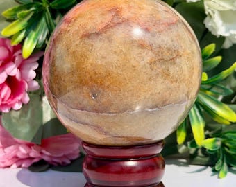 Puede incluir: Una esfera de piedra pulida que presenta una mezcla cálida y moteada de tonos bronceado, beige y marrón rojizo con vetas naturales visibles. La esfera descansa sobre un soporte de madera oscuro, circular y pulido con una base redondeada.