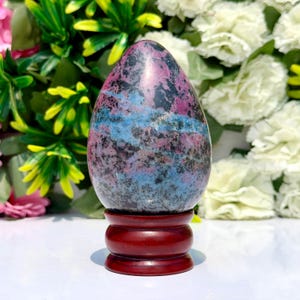 Op de afbeelding: Een eivormig stenen beeldhouwwerk met een gevlekt patroon van diep magenta, helderblauwe en zwarte mineraalinsluitingen. De gepolijste steen is gemonteerd op een gladde, ronde voet van donker roodbruin hout.