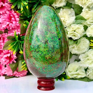 Puede incluir: Una escultura de piedra pulida en forma de huevo que presenta una base verde vibrante con inclusiones moteadas de color magenta y rojo oscuro. El objeto liso y redondeado está montado sobre una pequeña base de pedestal de madera marrón oscuro.