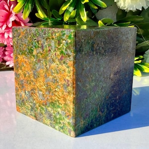 Op de afbeelding: Een vierkante decoratieve steen met een gevlekt patroon van groen, oranje en grijs. De stenen kubus heeft een gepolijst oppervlak en een natuurlijk, aards uiterlijk. De kleuren en texturen creëren een unieke, artistieke esthetiek.