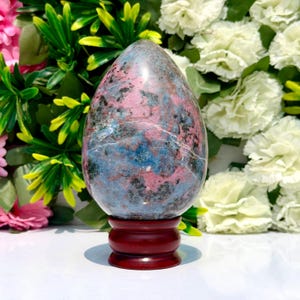 Puede incluir: Una escultura de piedra pulida en forma de huevo con un patrón moteado de inclusiones minerales rosas, azules y gris oscuro. La piedra se exhibe en posición vertical sobre un soporte de madera marrón oscuro con una base escalonada.