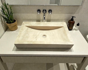May include: A rectangular, light-colored stone sink with a curved indentation for water flow. The sink is set on a white vanity with a drawer. A potted plant, mirror, and soap dispensers are visible in the background.