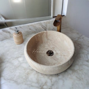 Op de afbeelding: Een ronde, beige stenen waskom met een afvoergat in het midden. De waskom staat op een wit marmeren aanrechtblad. Een gouden kraan is zichtbaar op de achtergrond.