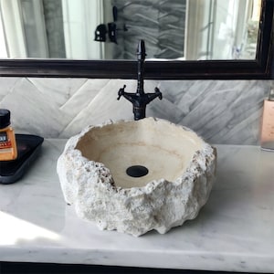 May include: A round vessel sink carved from light beige natural stone with a rough, textured exterior and a smooth interior basin. It features a dark matte black metal tap with two handles and a matching black circular drain plug.