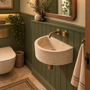 May include: A wall-mounted semi-circular beige travertine stone sink featuring a brushed brass faucet and handle. The sink is installed against a sage green vertical paneled wall, complemented by a matching brass drain pipe underneath.