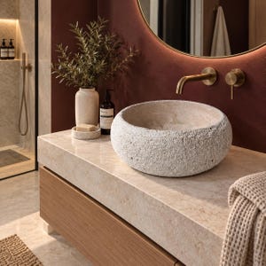 May include: A round, textured, off-white stone vessel sink sits on a light beige marble countertop. Beside it is a matching stone soap dish with a bar of soap, a tall speckled vase with greenery, and a dark amber bottle with a black pump.
