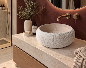 May include: A round, textured, off-white stone vessel sink sits on a light beige marble countertop. Beside it is a matching stone soap dish with a bar of soap, a tall speckled vase with greenery, and a dark amber bottle with a black pump.