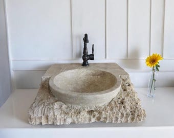 May include: A rustic stone sink with a rough, textured surface and a round basin. The sink is set on a white countertop and features a black faucet with a decorative handle.