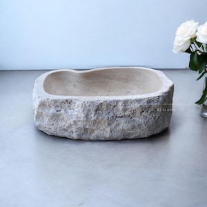 May include: A rectangular stone vessel sink with a rough, natural exterior texture and a smooth, light beige interior basin. The sink features a porous, organic rock surface and a wide, shallow bowl shape, presented with a white rose nearby.