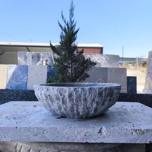 Op de afbeelding: Een getextureerde, grijze stenen kom met een ruwe buitenkant staat op een bijpassend stenen oppervlak. Achter de kom staat een kleine groenblijvende boom, met een heldere blauwe lucht op de achtergrond. De kom is een decoratief item.