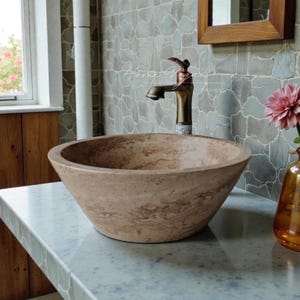 Op de afbeelding: Een ronde waskom van beige steen met natuurlijke aders en een gladde, taps toelopende buitenkant. De waskom staat onder een bronskleurige kraan. De tekst "Home Vogue DESIGNS" is zichtbaar op de muur achter de kraan.