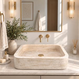 May include: A square beige stone vessel sink with a textured surface and a matching circular drain cover. The sink is mounted on a countertop with a wall-mounted gold tap, two vertical glass wall sconces, and a gold-framed rectangular mirror.
