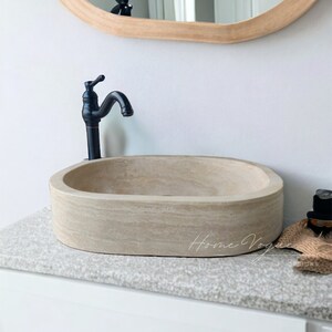 May include: An oval-shaped beige stone vessel sink with a natural textured finish, paired with a tall matte black tap. The sink is positioned on a speckled countertop, and the text Home Vogue Designs is visible on the lower right side.