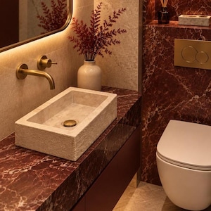 May include: A rectangular stone vessel sink with a brass drain sits on a deep red marble countertop. A wall-mounted brass tap, a white vase with red foliage, a mirror, a white wall-hung toilet, and a brass flush plate are present.