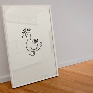 May include: White framed art print featuring a black line drawing of a duck with the words "WHAT THE" above it. The artwork is displayed on a white background and leans against a light gray wall.