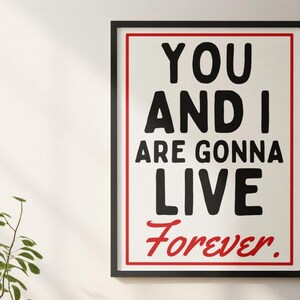 May include: A framed print with the words "YOU AND I ARE GONNA LIVE Forever." The text is black, with "Forever" in red. The print is in a black frame with a red border, and is mounted on a white wall. A small plant is visible.