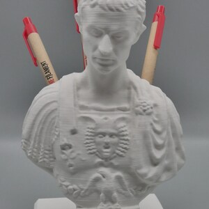 K&ouml;nnte beinhalten: Wei&szlig;e 3D-gedruckte B&uuml;ste einer Person mit einem detaillierten Gesicht und einem dekorativen Brustst&uuml;ck. Drei rote Stifte sind in den Kopf der B&uuml;ste gesteckt, wobei der Text "The Filament" auf einem der Stifte sichtbar ist.