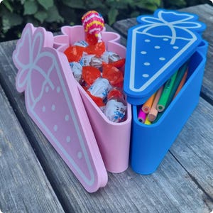 Könnte beinhalten: Zwei erdbeerförmige Behälter, einer rosa und einer blau, stehen auf einer Holzoberfläche. Der rosa Behälter enthält verpackte Süßigkeiten und einen Lutscher. Der blaue Behälter enthält Buntstifte. Die Behälter haben einen Deckel mit Erdbeermuster.