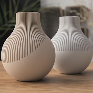 Könnte beinhalten: Zwei dekorative Vasen mit modernem Design. Eine Vase ist hellbeige, die andere weiß. Beide haben einen abgerundeten Boden und einen strukturierten, gerippten oberen Teil. Sie stehen auf einer Holzoberfläche.
