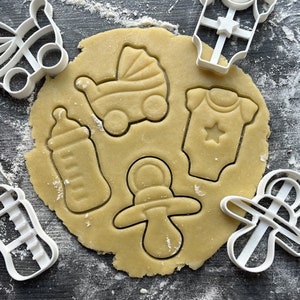 May include: A close-up of a cookie sheet with various baby-themed cookie cutters. The cutters are for a baby bottle, a pacifier, a baby onesie, a baby carriage, and a baby rattle.