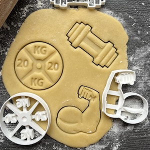 Könnte beinhalten: Nahaufnahme eines Keksausstecher-Sets zum Backen. Das Set enthält eine Hantel, einen Bizeps und eine Gewichtsplatte mit der Aufschrift "20 KG 20".
