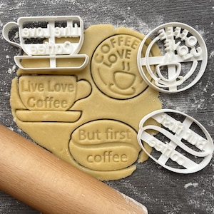 Op de afbeelding: Witte plastic koekjessnijders in verschillende vormen, waaronder een koffiekopje met de tekst "Live Love Coffee", een koffieboonvorm met "But first coffee" en een cirkelvormig ontwerp met "Coffee Love". De snijders liggen op uitgerold deeg.