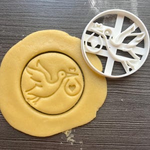 Op de afbeelding: Een witte plastic koekjesvormer in de vorm van een ooievaar die een baby in een tas draagt. De vormer ligt op een houten oppervlak naast een koekdeeg dat met de vormer is uitgestoken.