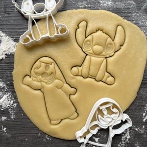 Op de afbeelding: Drie koekjesvormen, één in de vorm van Stitch uit Lilo & Stitch, één in de vorm van een meisje met een jurk en één in de vorm van een hond. De vormen liggen op een vel koekdeeg.