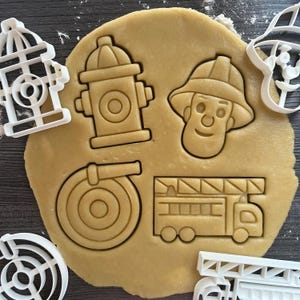 Peut inclure: Une pâte à biscuits avec des découpes d'une bouche d'incendie, du visage d'un pompier, d'un tuyau d'incendie et d'un camion de pompiers. Des emporte-pièces en plastique blanc d'une bouche d'incendie, d'un chien et d'une échelle sont autour de la pâte.