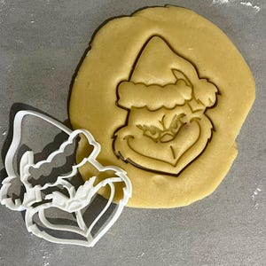 Könnte beinhalten: Ein weißer Ausstecher in Form des Gesichts und des Huts des Grinch, neben einem Keksteigausschnitt desselben Designs. Der Keksteig ist hellbraun.