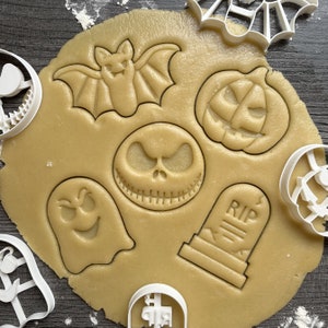Könnte beinhalten: Nahaufnahme eines Backblechs mit verschiedenen Halloween-förmigen Ausstechern. Die Ausstecher beinhalten eine Fledermaus, einen Kürbis, ein Gespenst, einen Grabstein mit der Aufschrift "RIP" und einen Schädel mit einem genähten Mund. Die Ausstecher befinden sich auf einer hellbraunen Oberfläche.