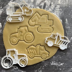 May include: A selection of white plastic cookie cutters in the shapes of construction vehicles, including a tractor, dumper truck, and excavator, pressed into a round of light brown dough. The cutters are arranged around the dough.