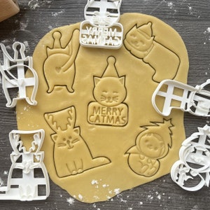 Peut inclure: Un ensemble de neuf emporte-pièces en plastique blanc en forme de chats, de rennes et de bas de Noël. Les emporte-pièces sont disposés sur une surface brune avec une pâte étalée. La pâte a été découpée avec les emporte-pièces pour créer des formes de chats, de rennes et de bas. Les emporte-pièces portent l'inscription "Merry Catmas" sur l'un d'eux.