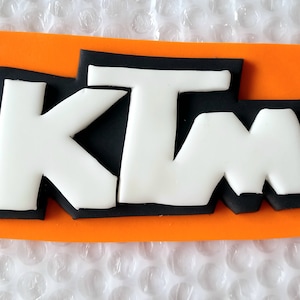 May include: A rectangular orange and black logo with the letters "KTM" in white. The logo is on a rectangular orange background. The letters are outlined in black.