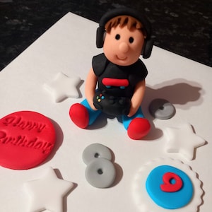 Puede incluir: Decoración de pastel con una figura de fondant de un jugador con auriculares y un mando. Rodeada de estrellas blancas, círculos grises, un círculo rojo con "Happy Birthday" y un círculo azul y blanco con el número 9.
