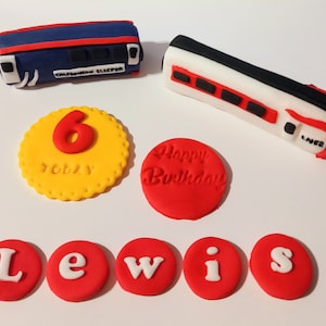 Op de afbeelding: Eetbare taartdecoraties met twee treinwagondesigns, één blauw met de tekst "CALEDONIAN SLEEPER" en één wit met de tekst "LNER". Ook inbegrepen zijn een gele cirkel met een rode "6" en de tekst "TODAY", een rode cirkel met de tekst "Happy Birthday" en rode letters die "LEWIS" spellen.