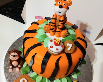 May include: A round orange cake decorated with black stripes, a tiger figurine, and a monkey figurine. The cake has green leaf accents, a cupcake, and a wooden number 7. The cake is in a white box.