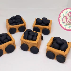 Può includere: Cinque decorazioni per cupcake in miniatura a forma di carrelli per il carbone. Ogni carrello è marrone chiaro con ruote nere e pieno di caramelle nere rotonde. L'immagine include un logo che dice "Cleo's cupcakes".