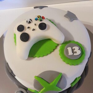 May include: A round cake decorated with an Xbox controller and accents. The cake is white with green and grey details, including a green scalloped edge with the number 13. The controller is white with black buttons and colourful accents.