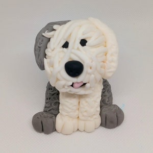 May include: A handmade Old English Sheepdog figurine. The dog is sitting, with a fluffy white and cream-coloured coat, grey ears and paws, and a black nose. The dog's tongue is pink.