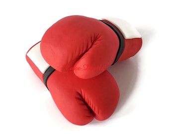 May include: Two red boxing gloves with white and black accents, arranged on a white background. The gloves are made of a smooth material and appear to be a decorative item. The text "Cake Devils" is visible on the gloves.