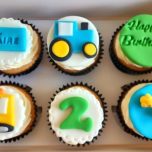 Pode incluir: Uma caixa de cupcakes decorados. Cada cupcake tem uma base de glacê branco com um forro preto. Os cupcakes são decorados com fondant azul, verde e amarelo. Alguns têm o texto "DÁIRE" e "Happy Birthday".