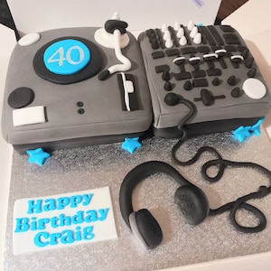 Puede incluir: Un pastel gris decorado como equipo de DJ, incluyendo un tocadiscos y una mesa de mezclas. El pastel tiene un círculo azul con el número 40, detalles de estrellas azules y un letrero rectangular que dice "Happy Birthday Craig" en texto azul.