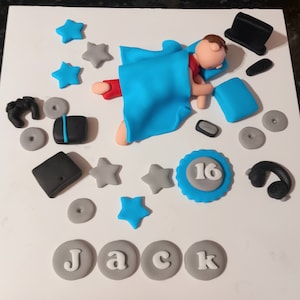 Könnte beinhalten: Ein Kuchen, der mit Fondantfiguren verziert ist. Eine Figur in einem blauen Umhang liegt, umgeben von Sternen, einem Laptop, einem Controller, Kopfhörern und dem Namen "JACK". Ein blau-weißes Emblem zeigt die Zahl "16".