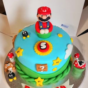Könnte beinhalten: Ein runder Kuchen, der mit einem blauen Himmel-Design verziert ist und eine Mario-Figur auf der Oberseite aufweist. Der Kuchen ist mit gelben Sternen, einem Fragezeichenblock und der Zahl 5 verziert. Zusätzliche Figuren und Pilze umgeben den Kuchen.