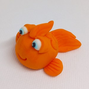 May include: A whimsical, orange goldfish figurine with large, expressive eyes and a smiling mouth. The fish has detailed fins and a rounded body, perfect for aquatic-themed decor or crafts.