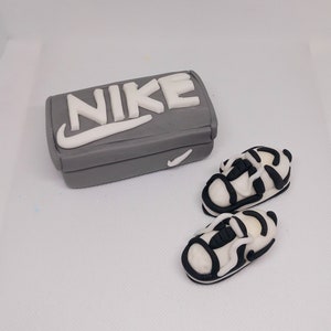 Peut inclure: Une boîte grise avec le mot "NIKE" en blanc et un logo swoosh blanc. Deux mini-baskets blanches et noires sont à côté de la boîte. La boîte et les chaussures sont sur une surface blanche.